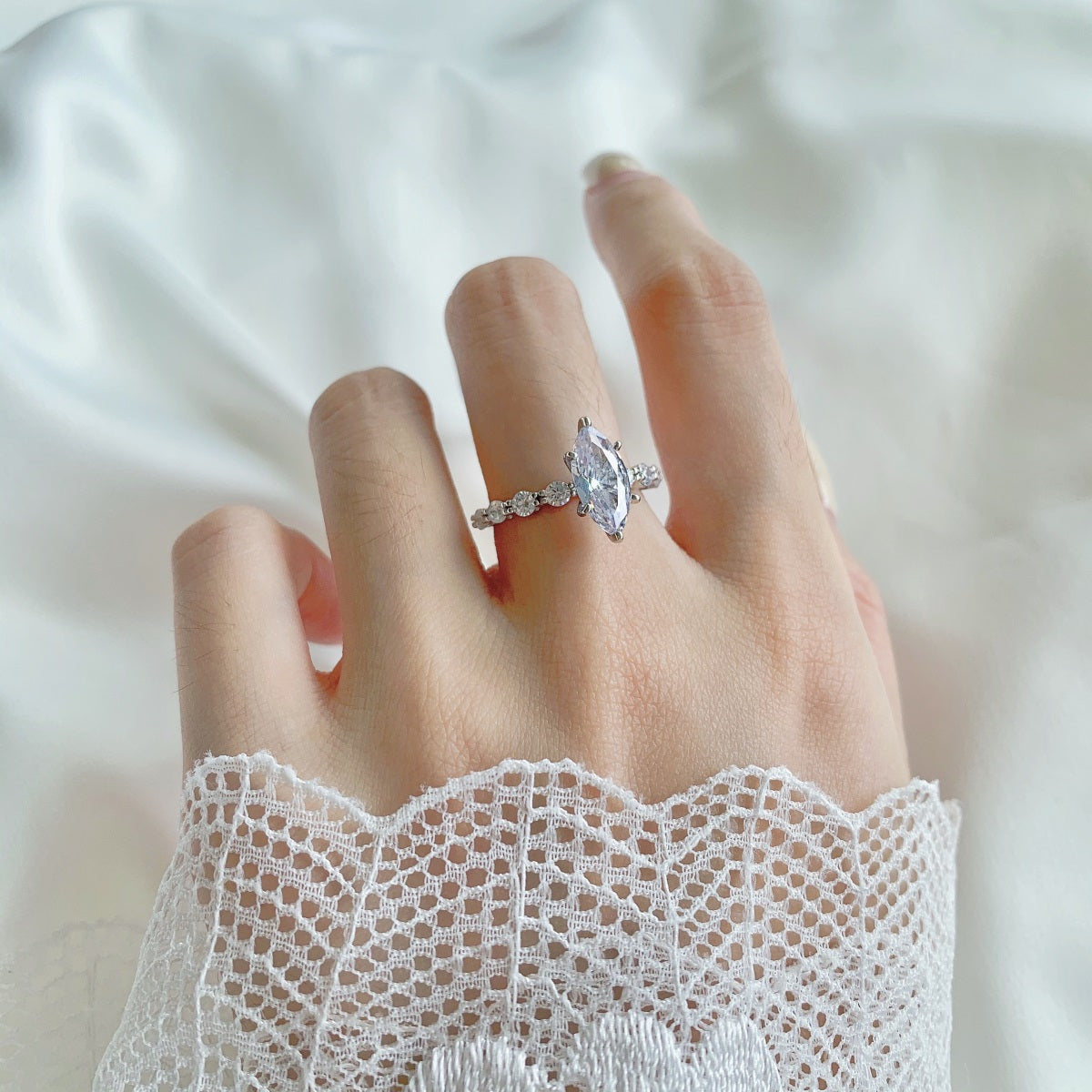 Hand wearing a silver marquise ring with a heart-shaped diamond on a white lace sleeve.