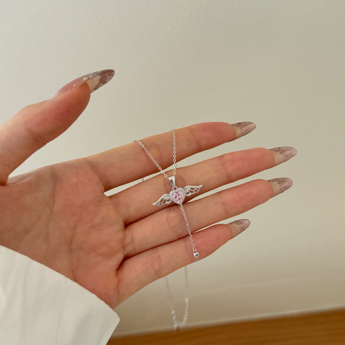 Hand holding a silver necklace with a pink heart pendant against a neutral background