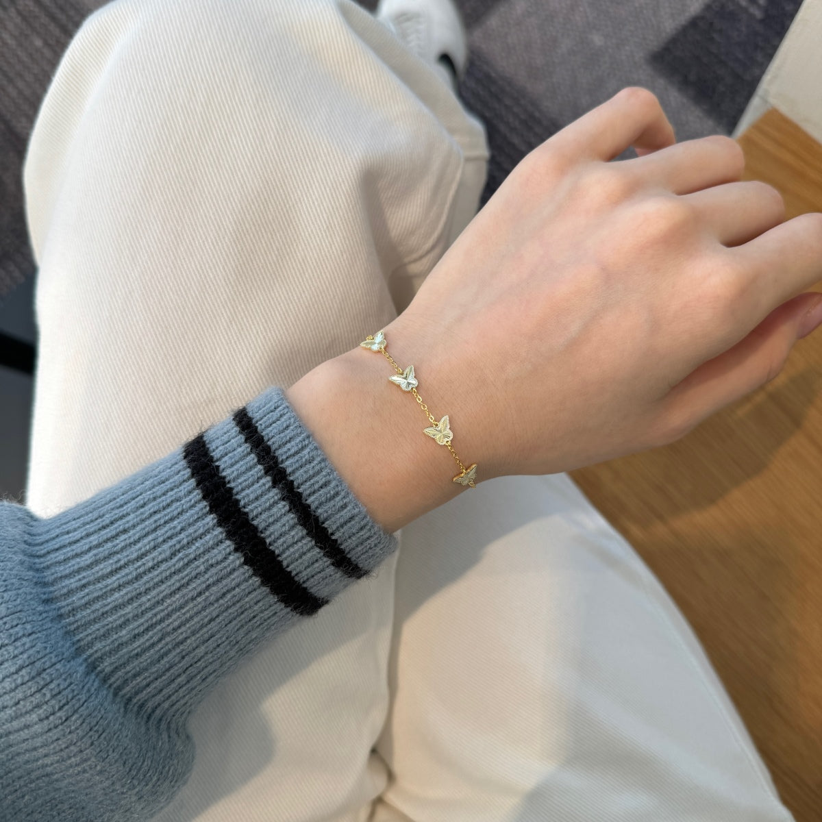 Hand wearing a gold bracelet with butterfly charms, wearing a blue sweater with black stripes.
