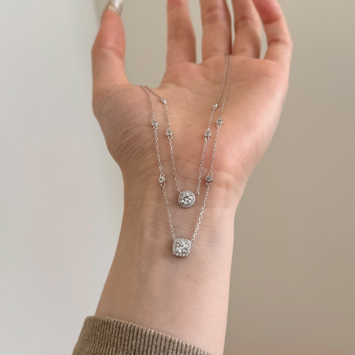 Hand wearing two silver necklaces with clear stones on a neutral background