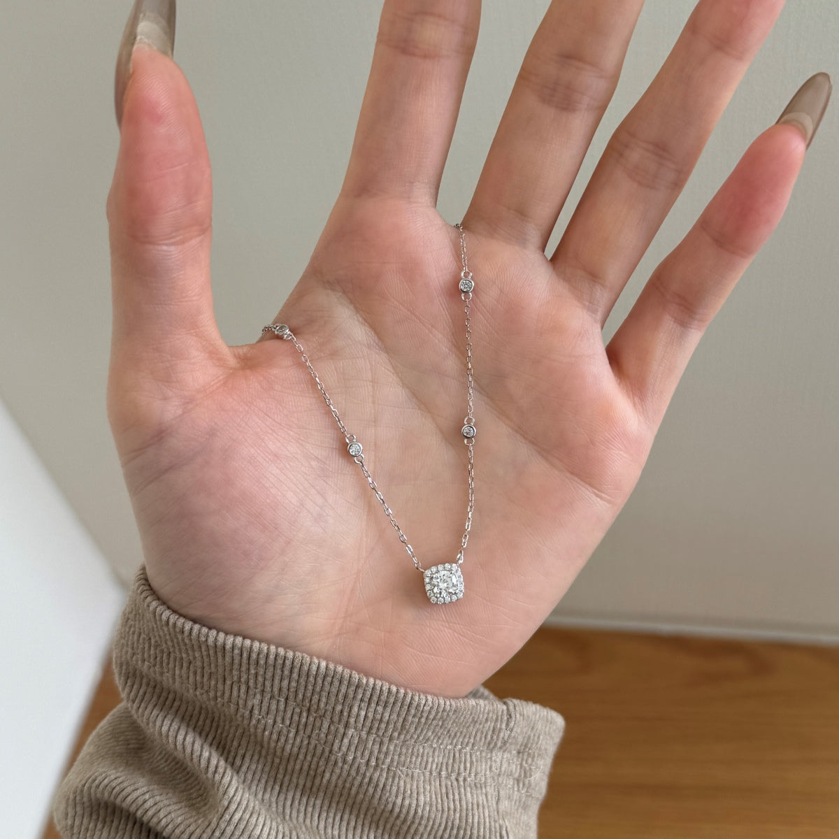 Hand wearing a silver necklace with a clear pendant against a neutral background