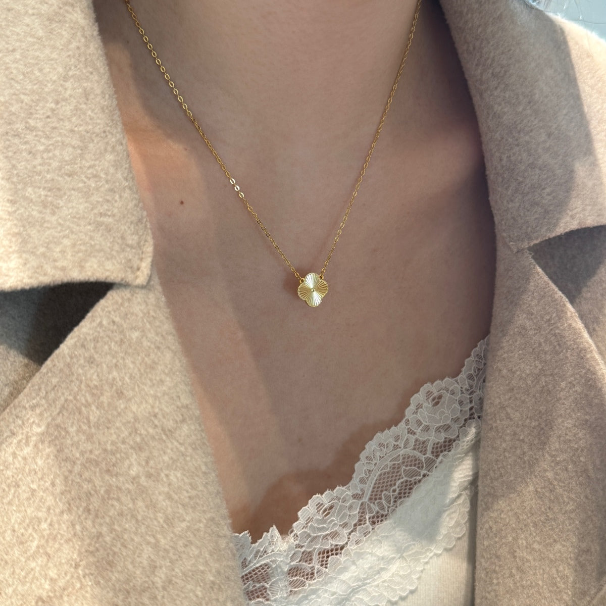 Gold necklace with a four leaf clover pendant worn by a person in a beige coat and lace top.
