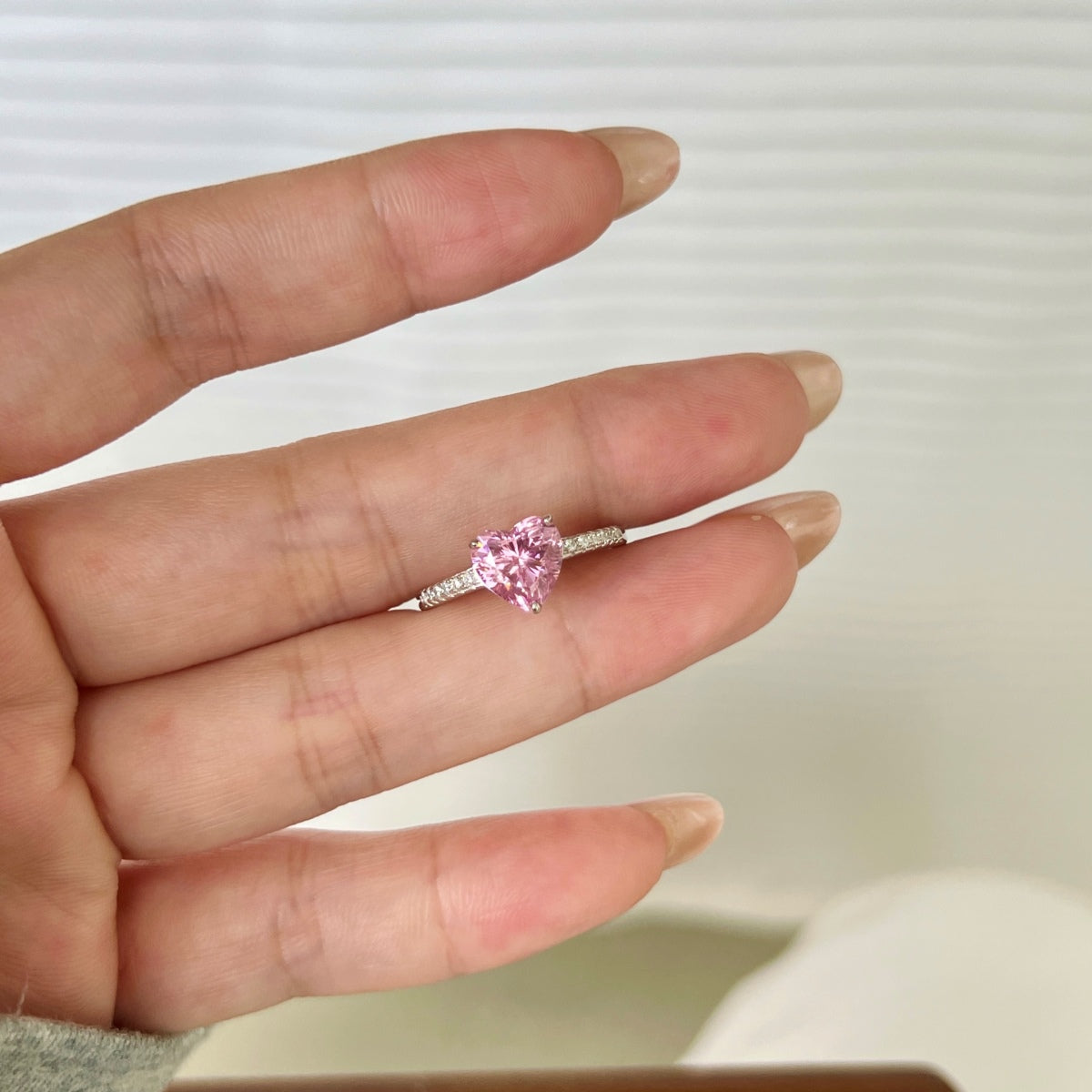 Hand wearing a ring with a pink heart-shaped gemstone on a neutral background