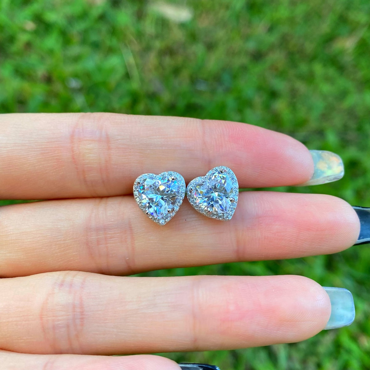 Heart-shaped diamond earrings held between fingers against a grassy background