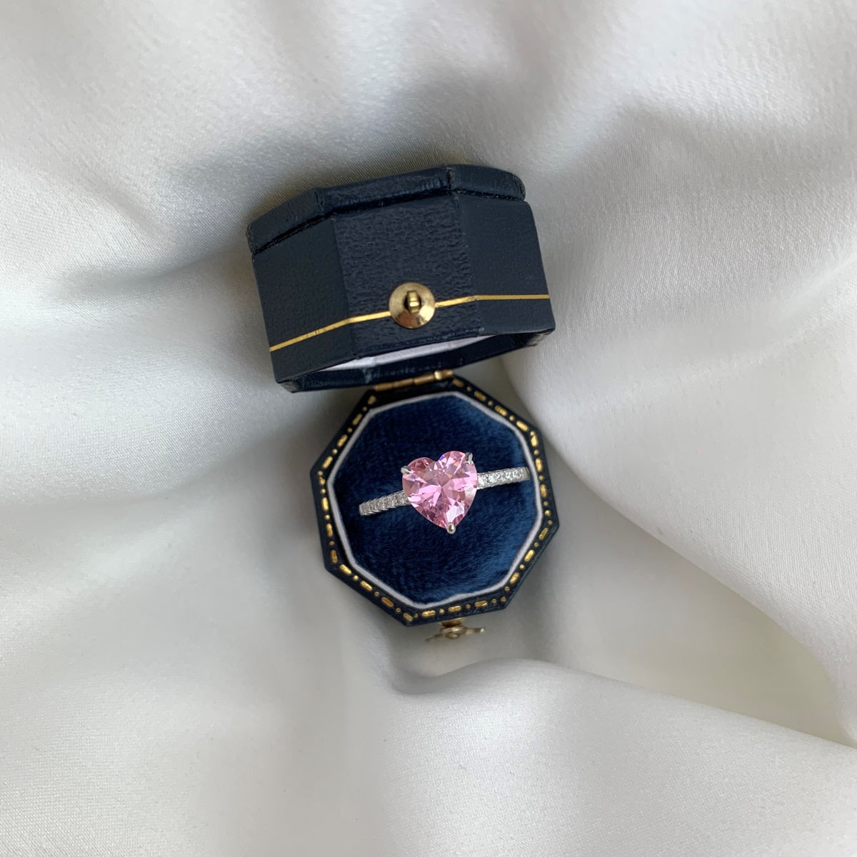 Heart-shaped pink gemstone ring in a blue jewelry box on a white fabric background