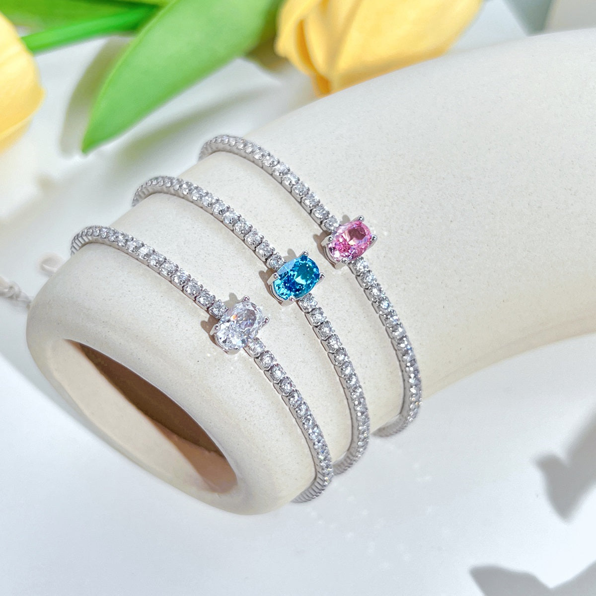 Three silver tennis bracelets with colorful gemstones on a white surface with yellow tulips in the background.