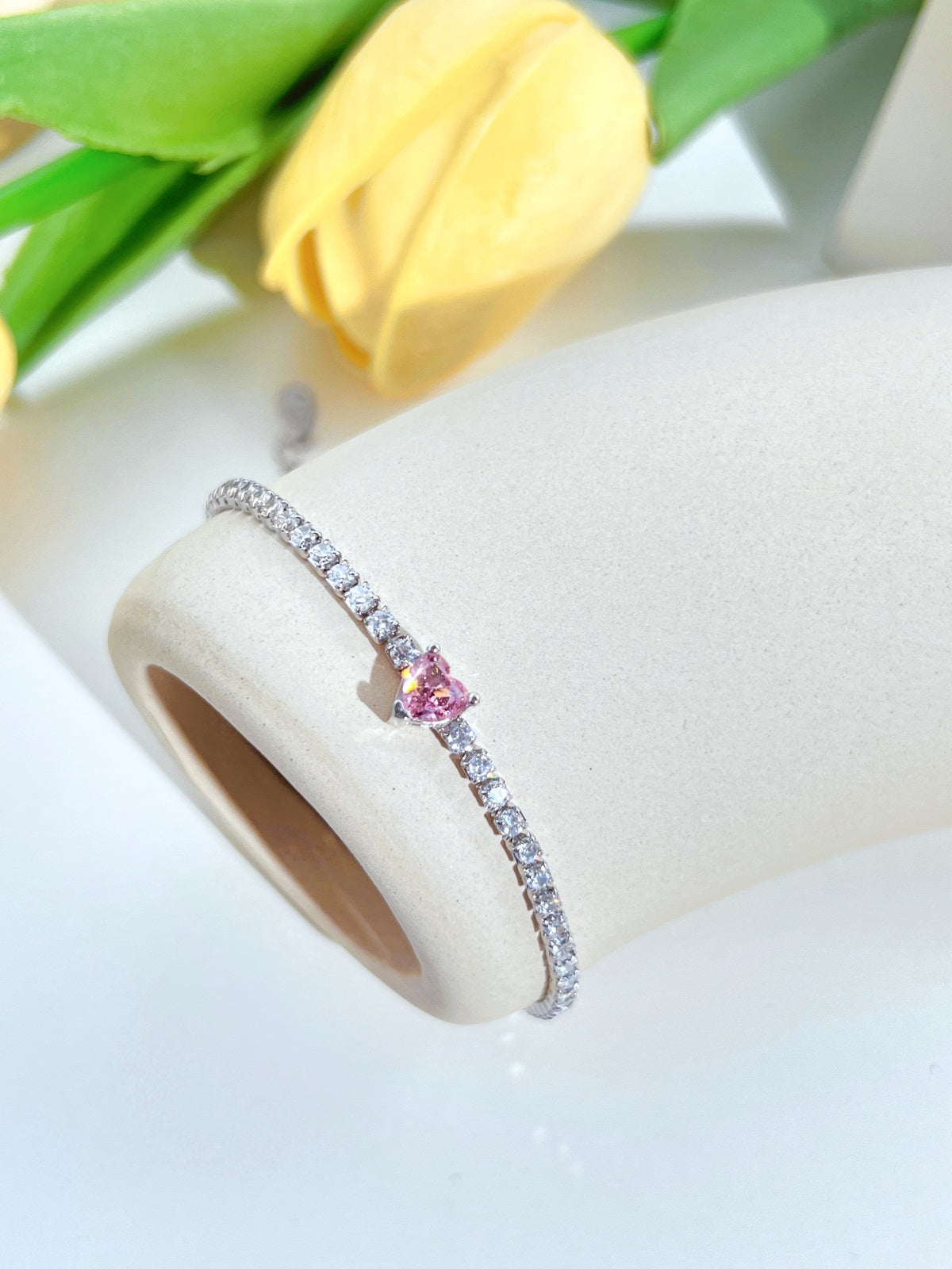 Silver tennis bracelet with a pink gemstone on a white surface with yellow tulips in the background