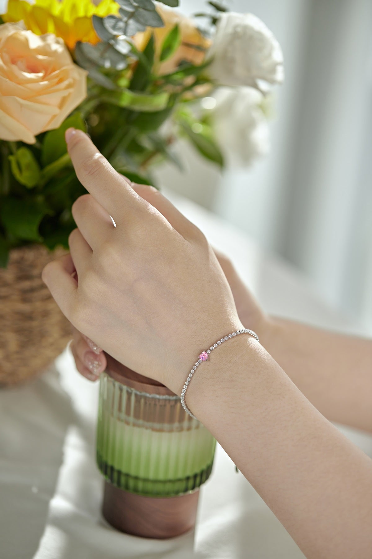 Close-up of a hand wearing a pink tennis bracelet with a blurred background of flowers and a vase.