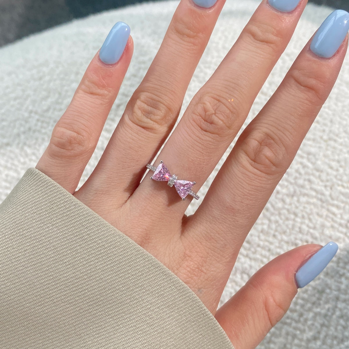 Hand wearing a pink bow-shaped ring with light blue nail polish on a neutral background