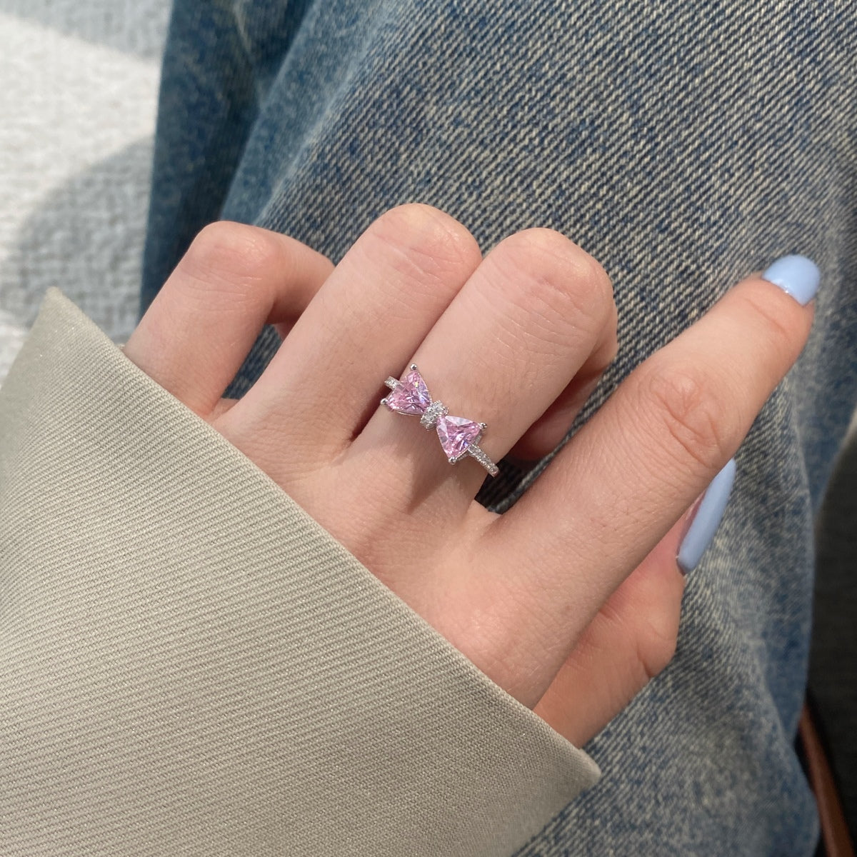 Hand wearing a pink gemstone ring with a bow design, against a denim background.